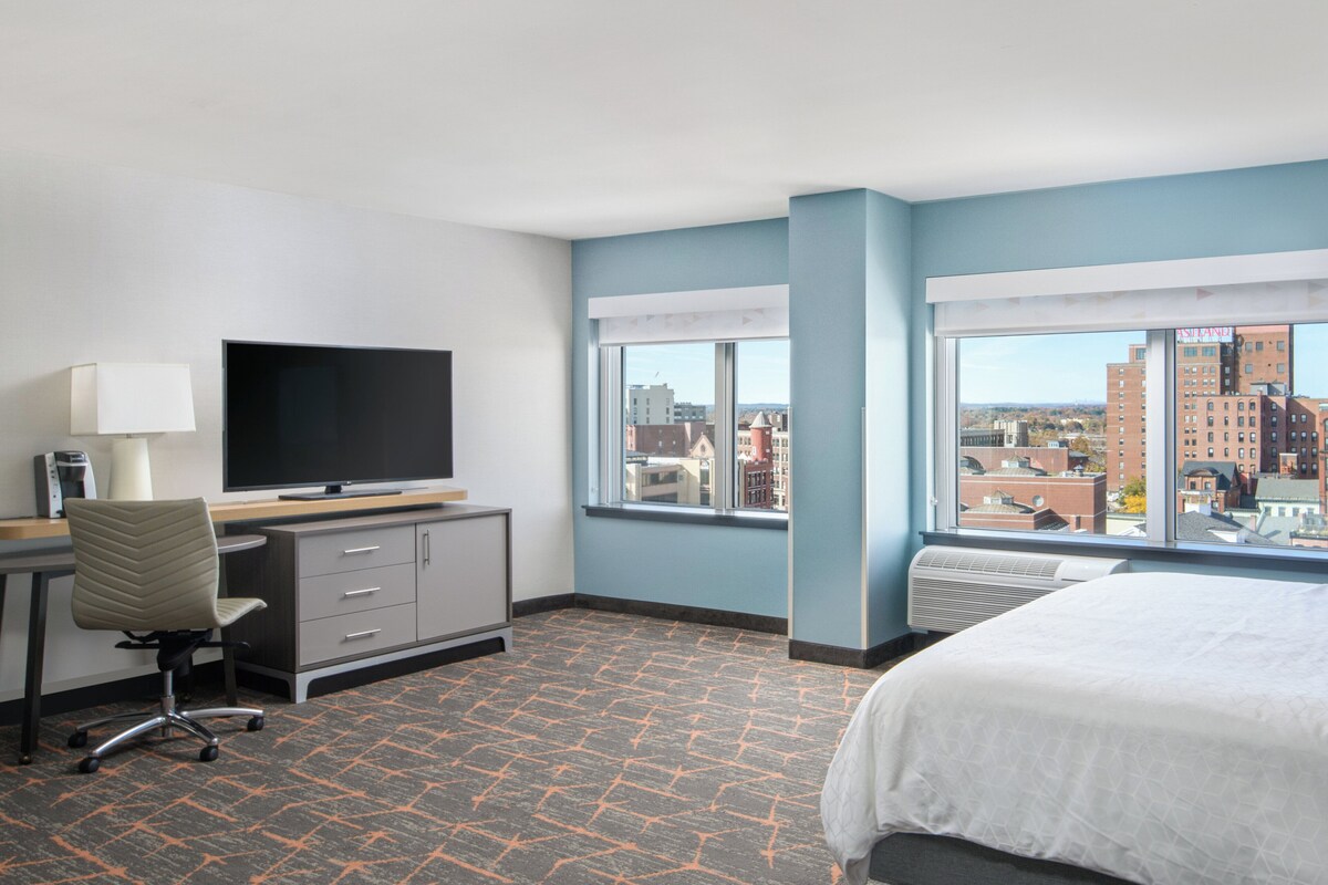 A hotel room with a bed, a desk with a TV, a chair, and a view of the city through large windows.