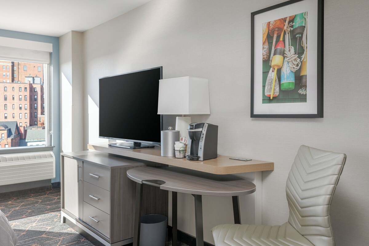 A hotel room with a flat-screen TV, a coffee maker, a framed artwork, and a chair.