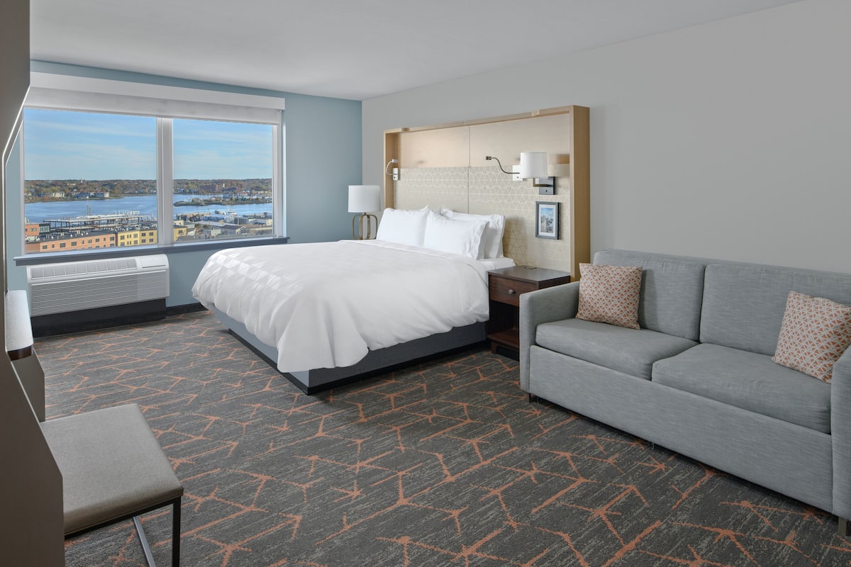 A hotel room with a large bed, a grey sofa, and a view of a cityscape through large windows.
