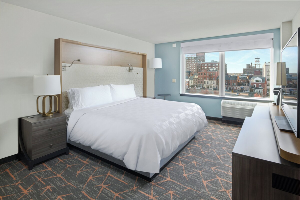 A hotel room with a large bed, a desk, a chair, a lamp, and a view of the city through a large window.