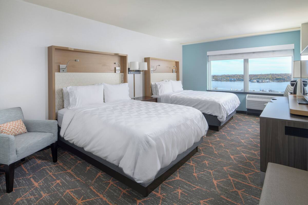 A hotel room with two beds, a desk, a chair, and a large window offering a view of the water.