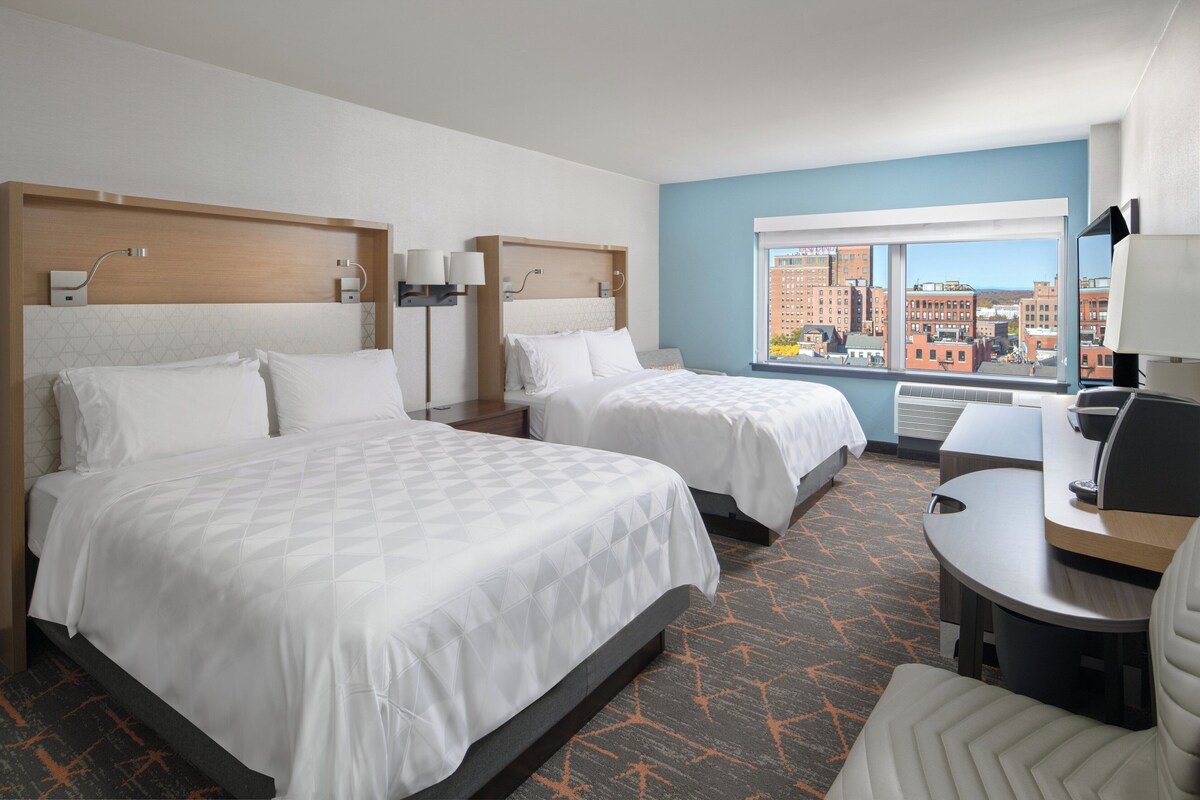 A hotel room with two beds, a desk, and a view of the city.