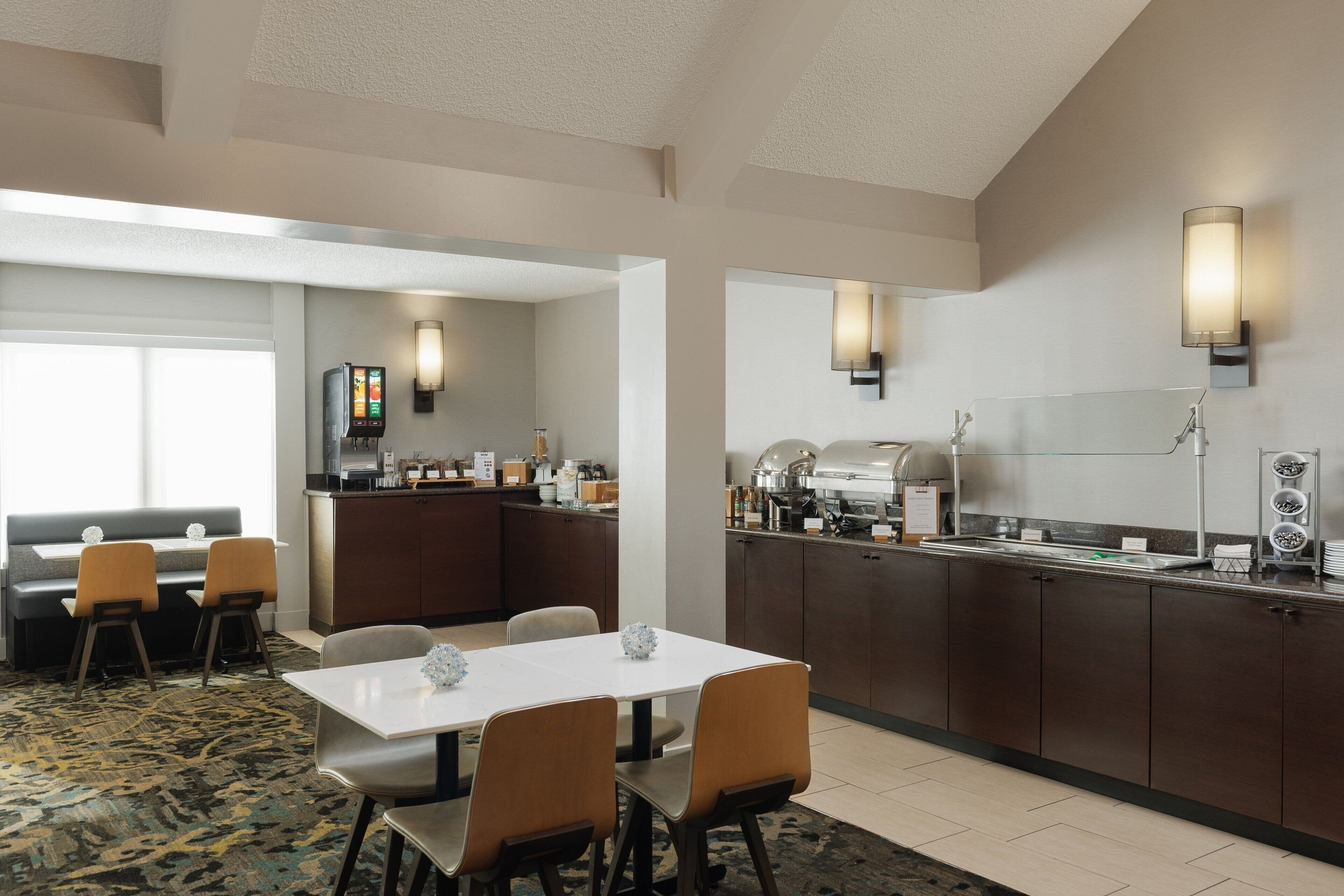 Comedor de hotel con barra libre, zona de estar con sofá y sillas, y cocina con estación de café.