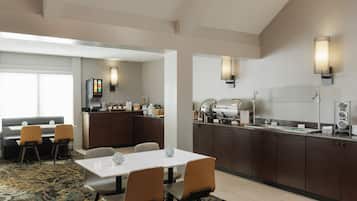Comedor de hotel con barra libre, zona de estar con sofá y sillas, y cocina con estación de café.
