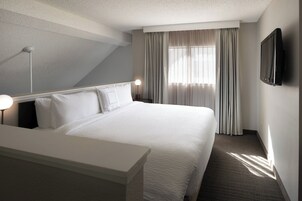 Habitación de hotel con una cama grande, una mesita de noche, un televisor de pantalla plana montado en la pared y una ventana con cortinas.