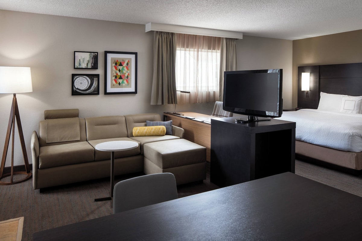 A hotel room with a sofa, a small table, a TV, a bed, and artwork on the wall.