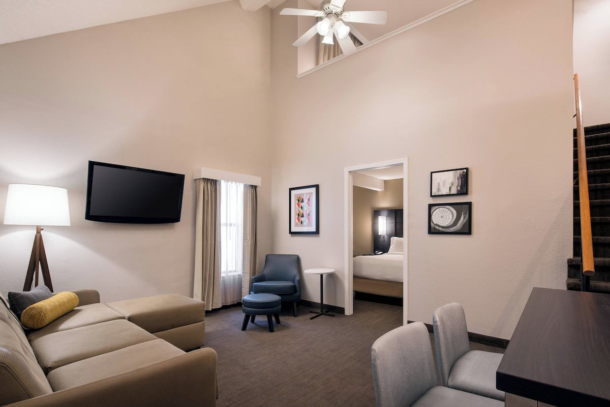 A hotel room with a sofa, armchair, ottoman, a small table, a TV, and a ceiling fan.
