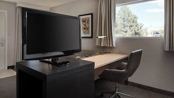 Hotelkamer met een groot flatscreen televisietoestel op een donker houten bureau, een lichtkleurig bureau, een moderne kantoorstoel, een wanddecoratie met een geometrisch kunstwerk en een raam met gordijnen.