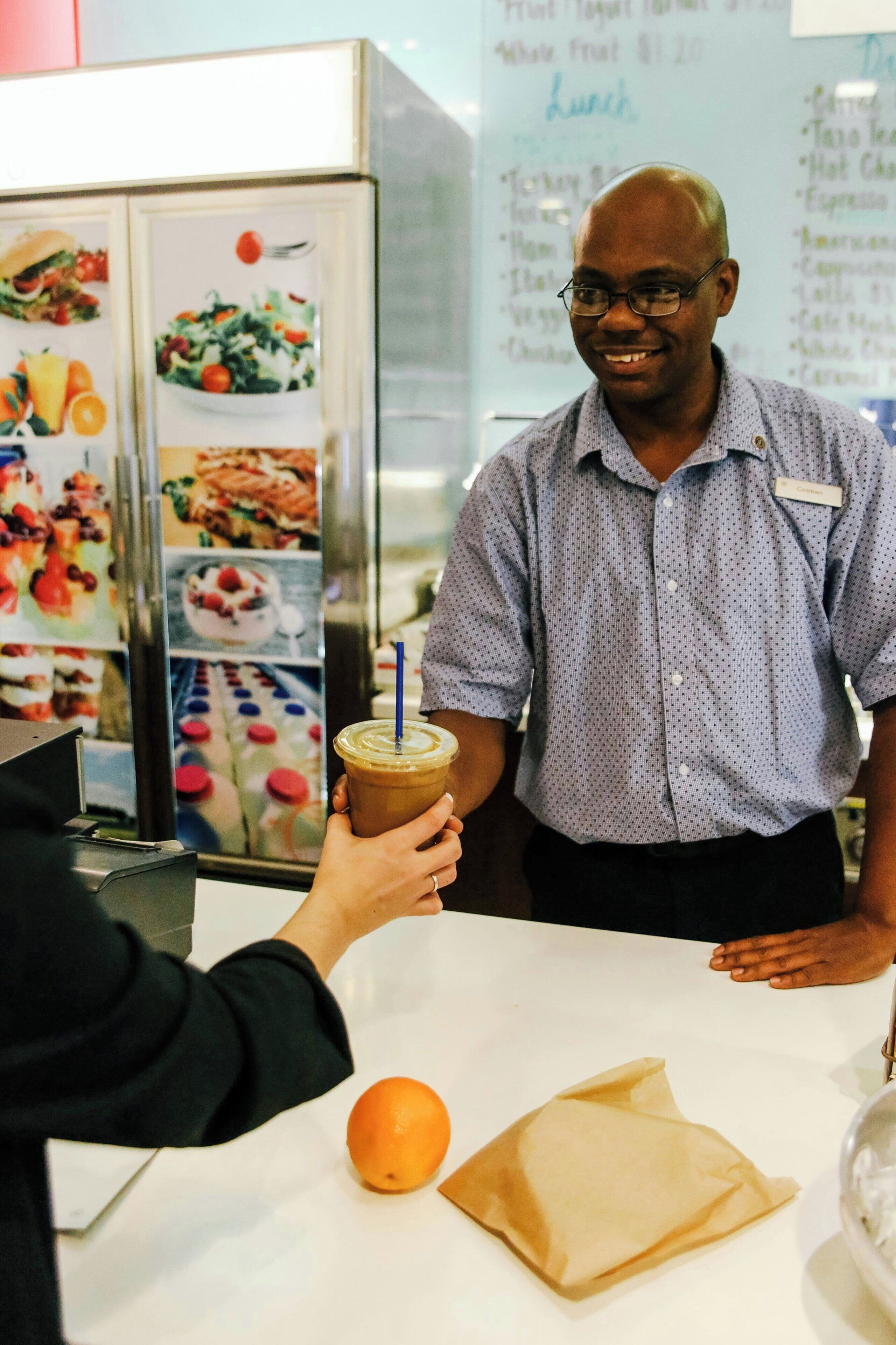 A person holding a drink with a straw, a menu board in the background, and a table with an orange and a napkin.