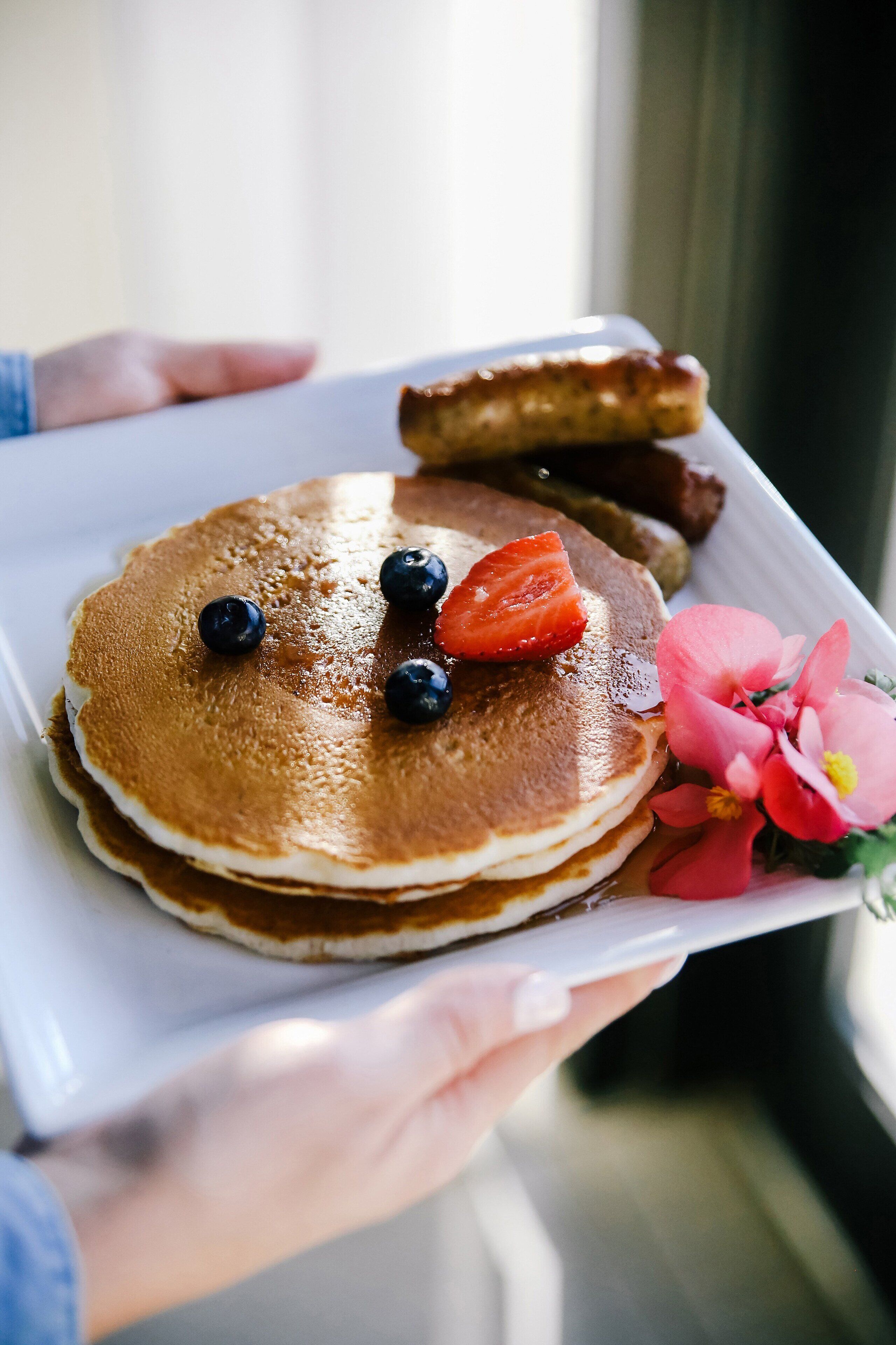 A person holding a tray with pancakes, blueberries, strawberries, and pink flowers.