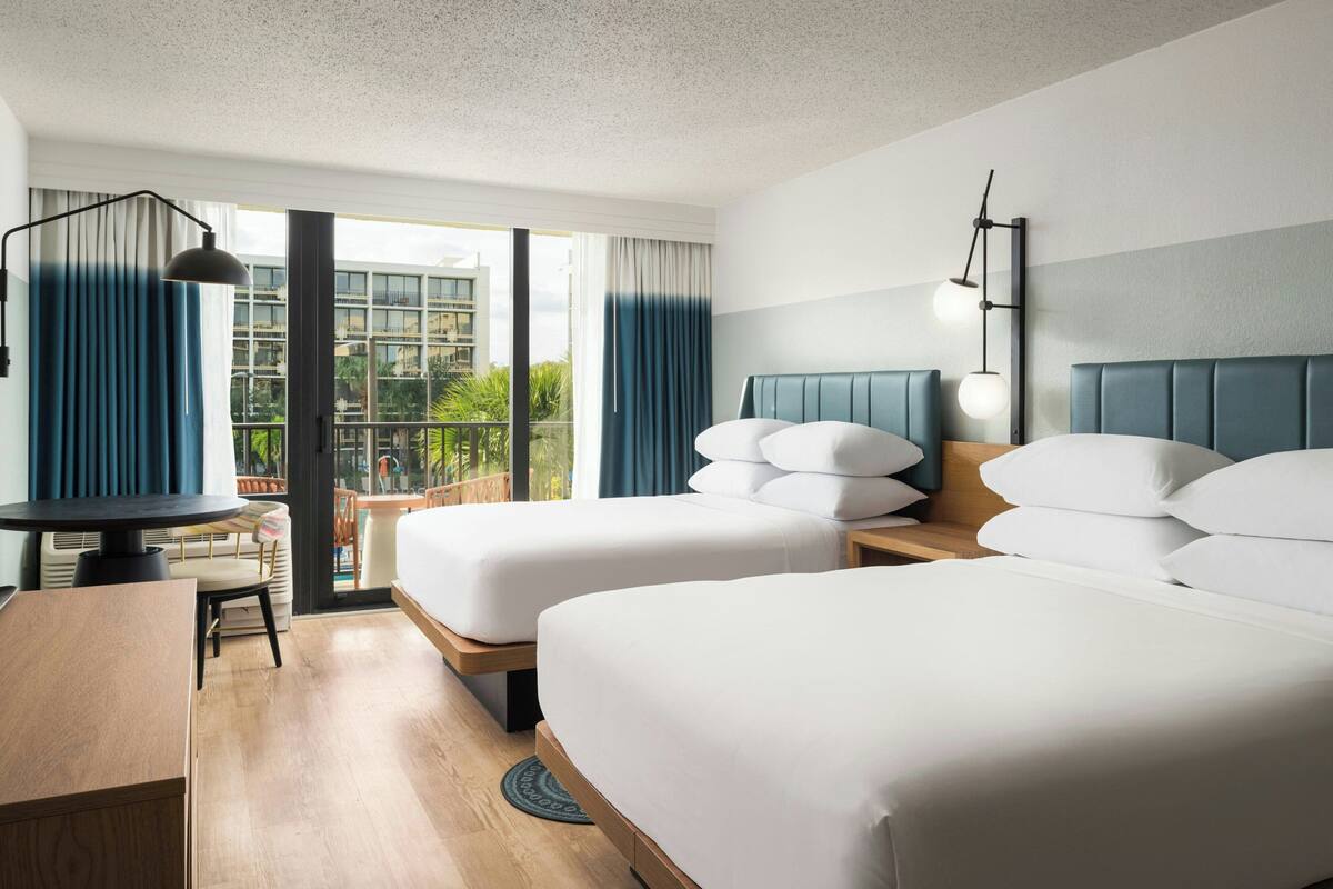 A hotel room with two beds, a desk, and a balcony with a view of buildings and greenery.