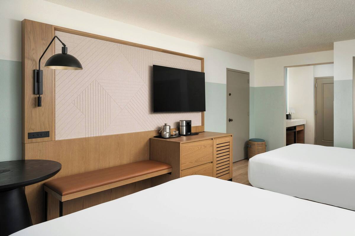 A hotel room with a flat-screen TV mounted on a wooden panel wall, a wooden desk with a bench, a bedside table, and a white bed.