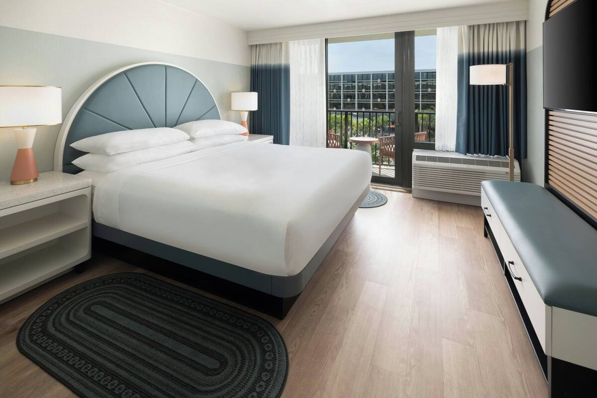 A hotel room with a large bed, bedside tables, a desk, a chair, a TV, and a view of the outdoors.