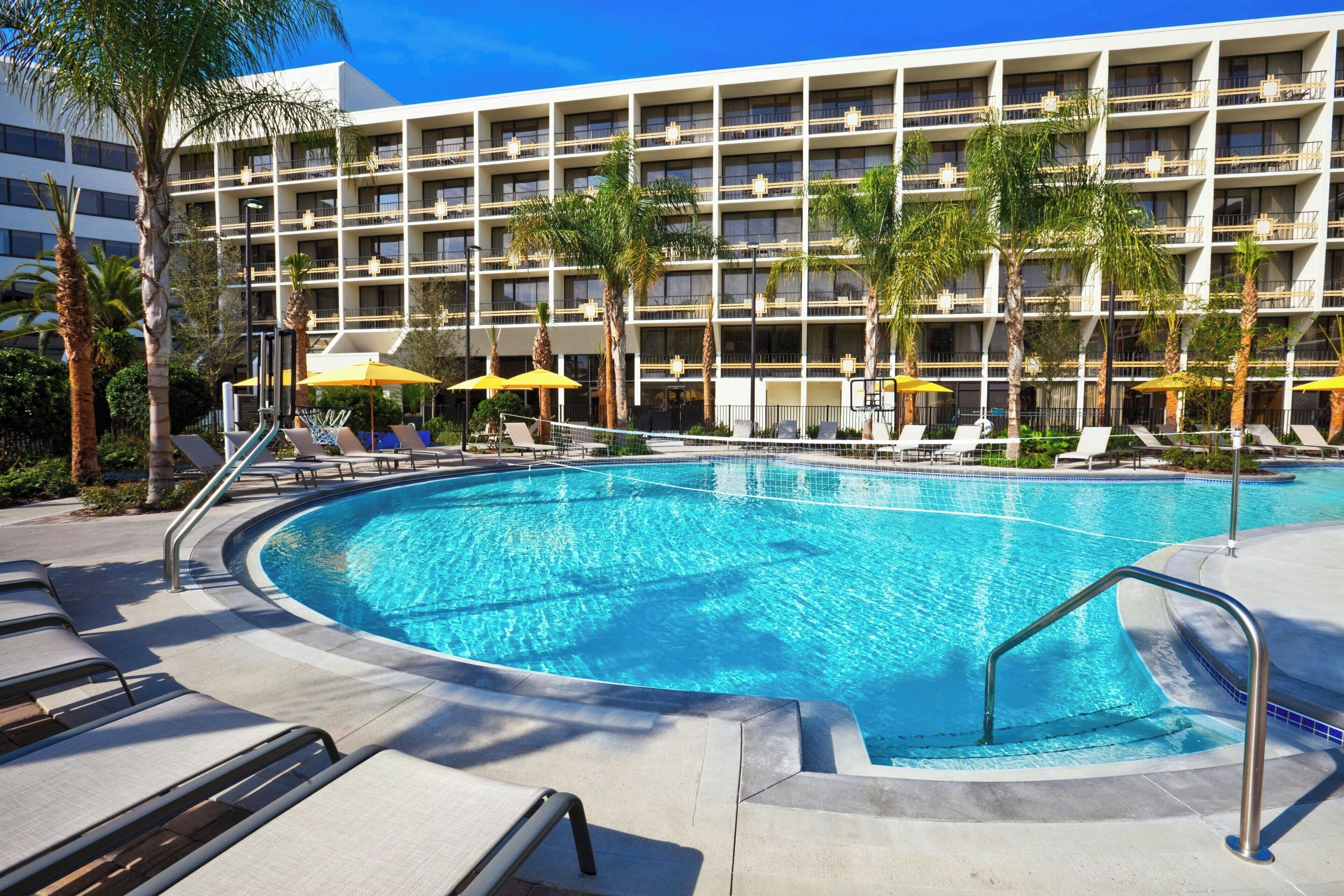 A hotel pool area with multiple lounge chairs and umbrellas, surrounded by palm trees and a multi-story building with balconies.