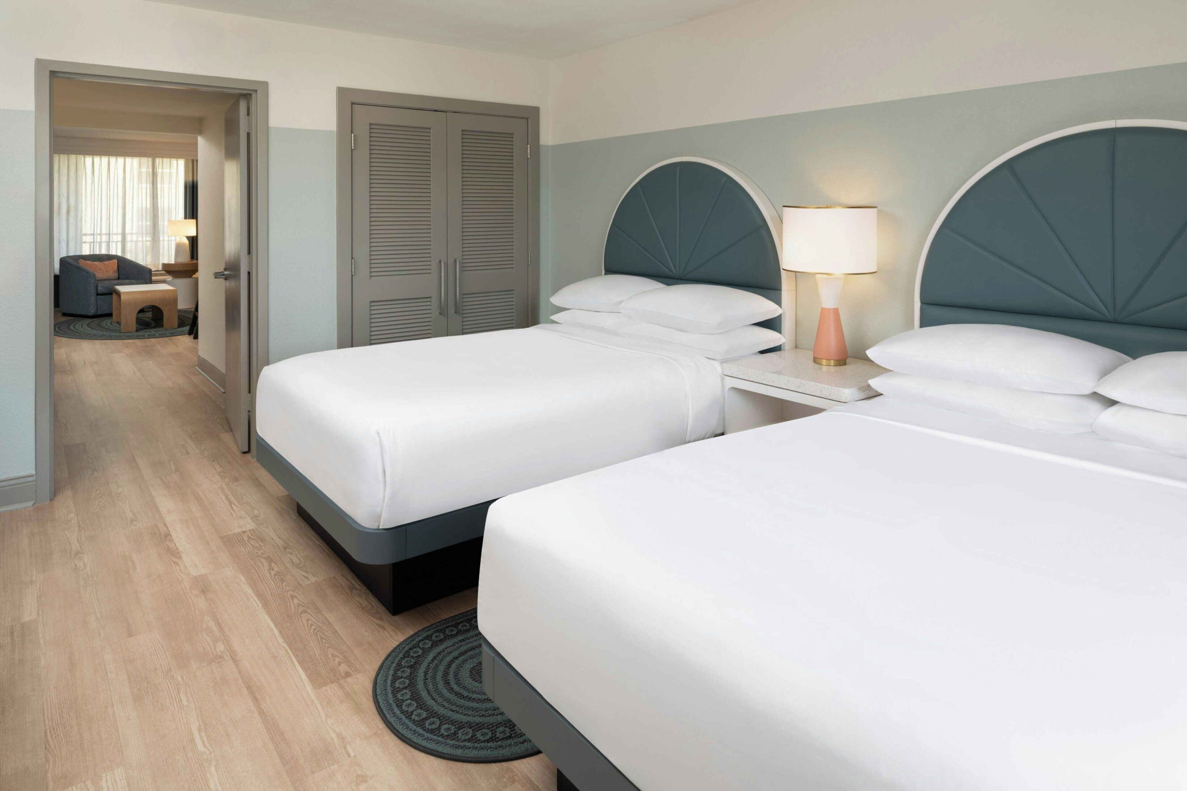 A hotel room with two beds, a desk with a lamp, a wardrobe, and a view of another room.