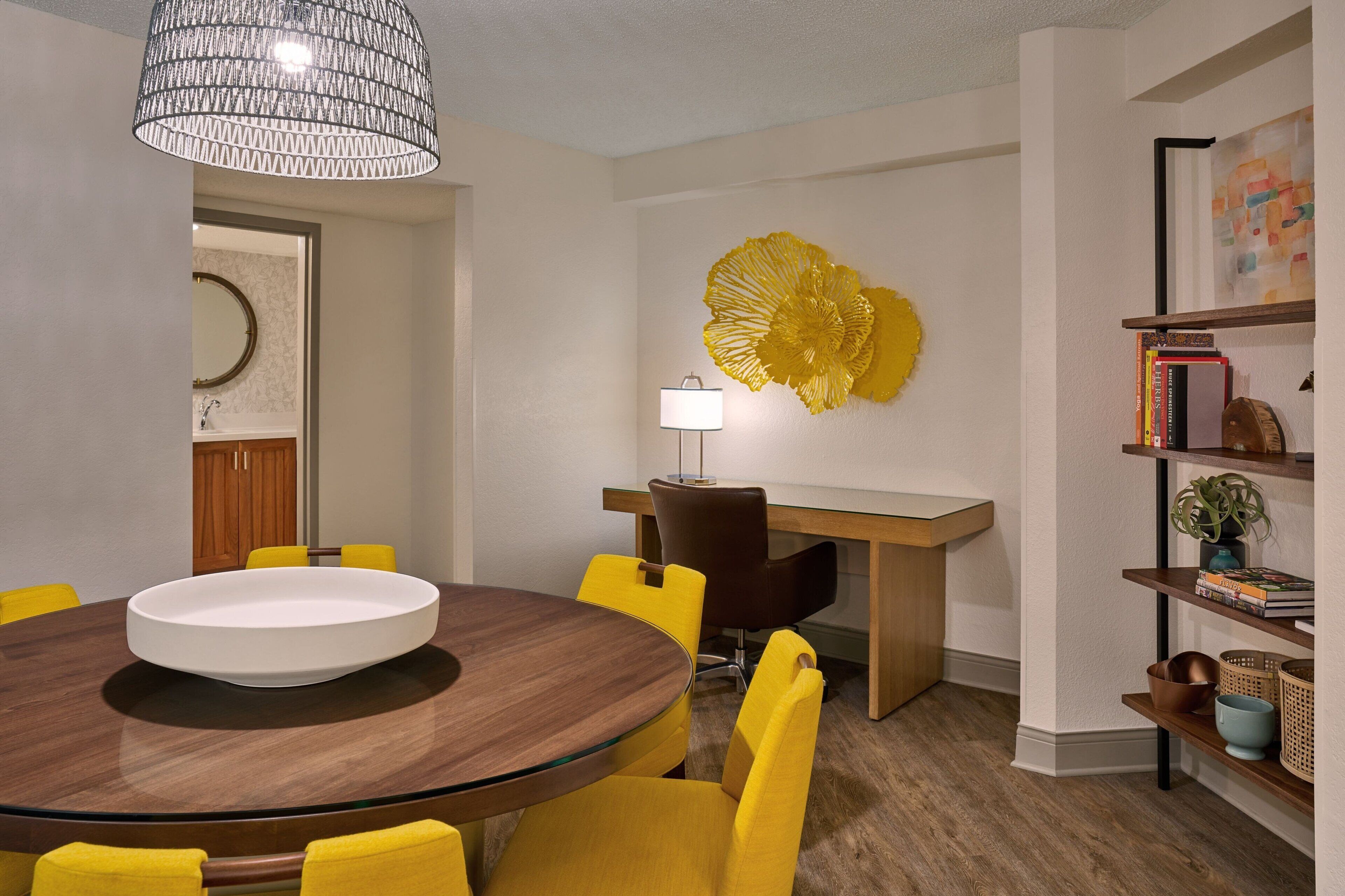 A dining area with a round table, yellow chairs, a wooden desk, a lamp, and a decorative yellow piece on the wall.