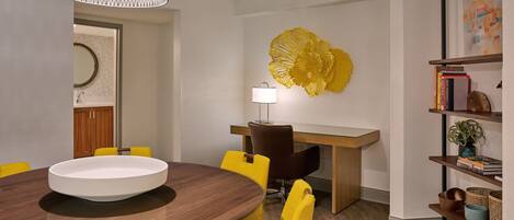 A dining area with a round table, yellow chairs, a wooden desk, a lamp, and a decorative yellow piece on the wall.