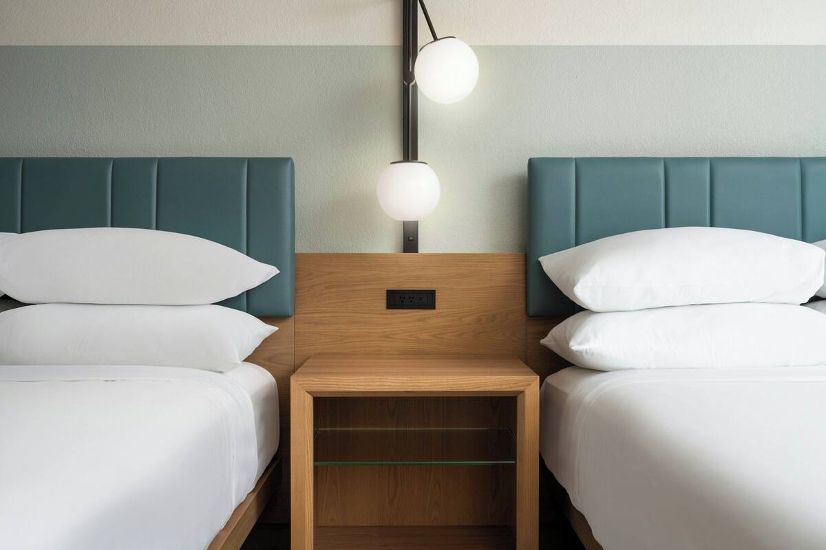 A hotel room with two beds, each with white bedding and teal headboards. There is a bedside table with a lamp between the beds.