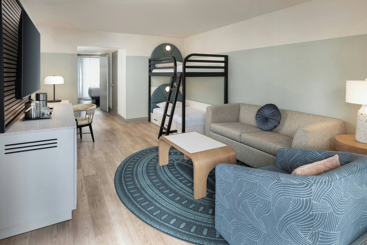 A compact hotel room with a bunk bed, a sofa, a small table, a desk, and a TV.