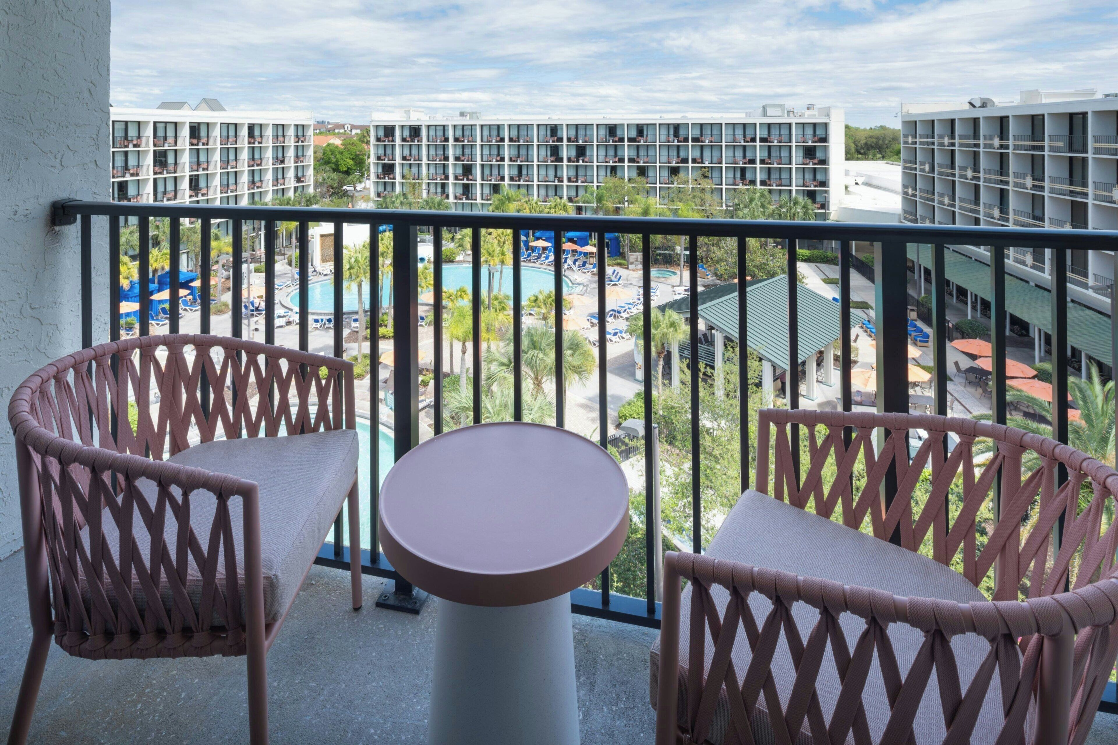 Un balcone con sedie in vimini e un tavolo, con vista su una zona piscina con ombrelloni e un edificio sullo sfondo.