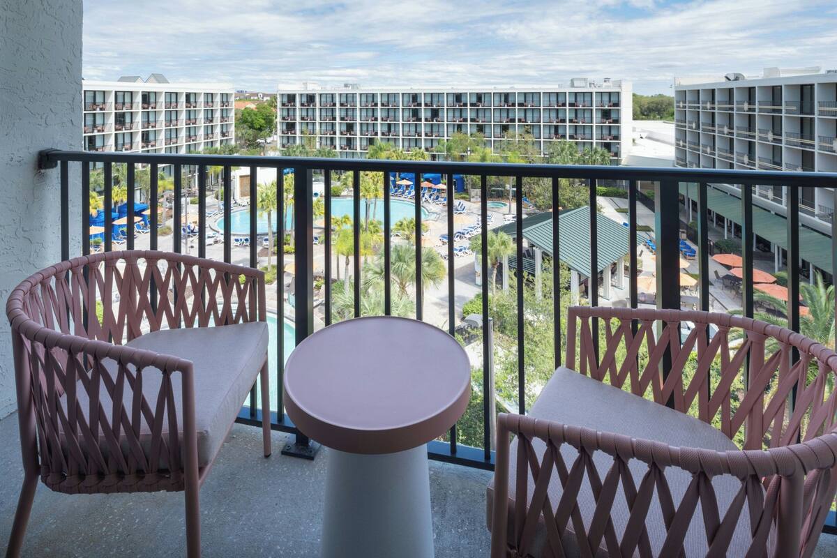 A balcony with wicker chairs and a table, overlooking a pool area with umbrellas and a building in the background.
