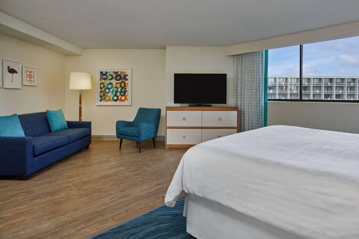 A hotel room with a bed, a sofa, a chair, a TV, and a window with a view of buildings.