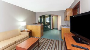 A hotel room with a sofa, a wooden table, a TV, and a kitchen area with a sink and cabinets.