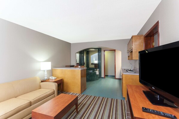A hotel room with a sofa, a wooden table, a TV, and a kitchen area with a sink and cabinets.
