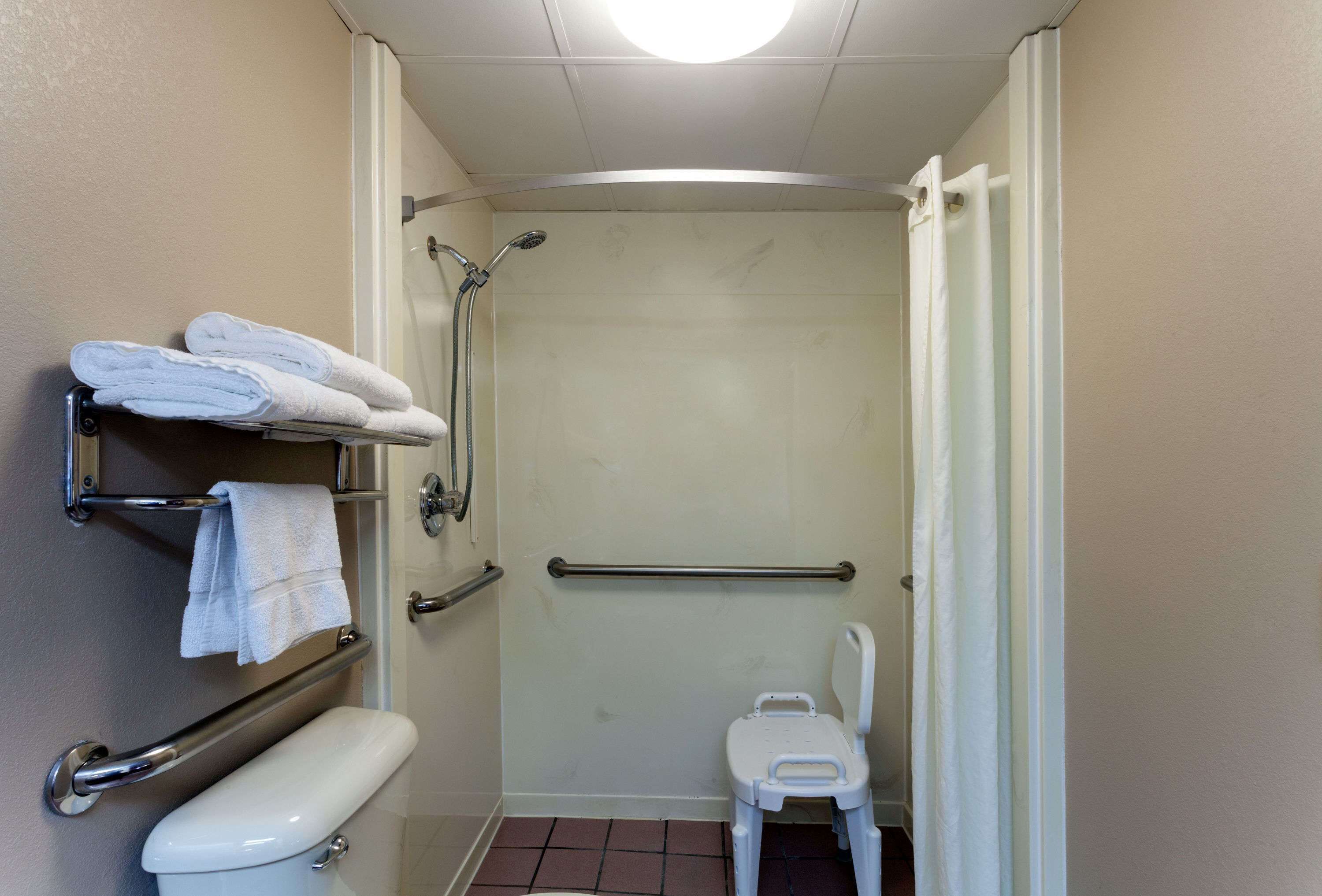 A small, accessible bathroom with a toilet, a shower with a grab bar, and a white chair.