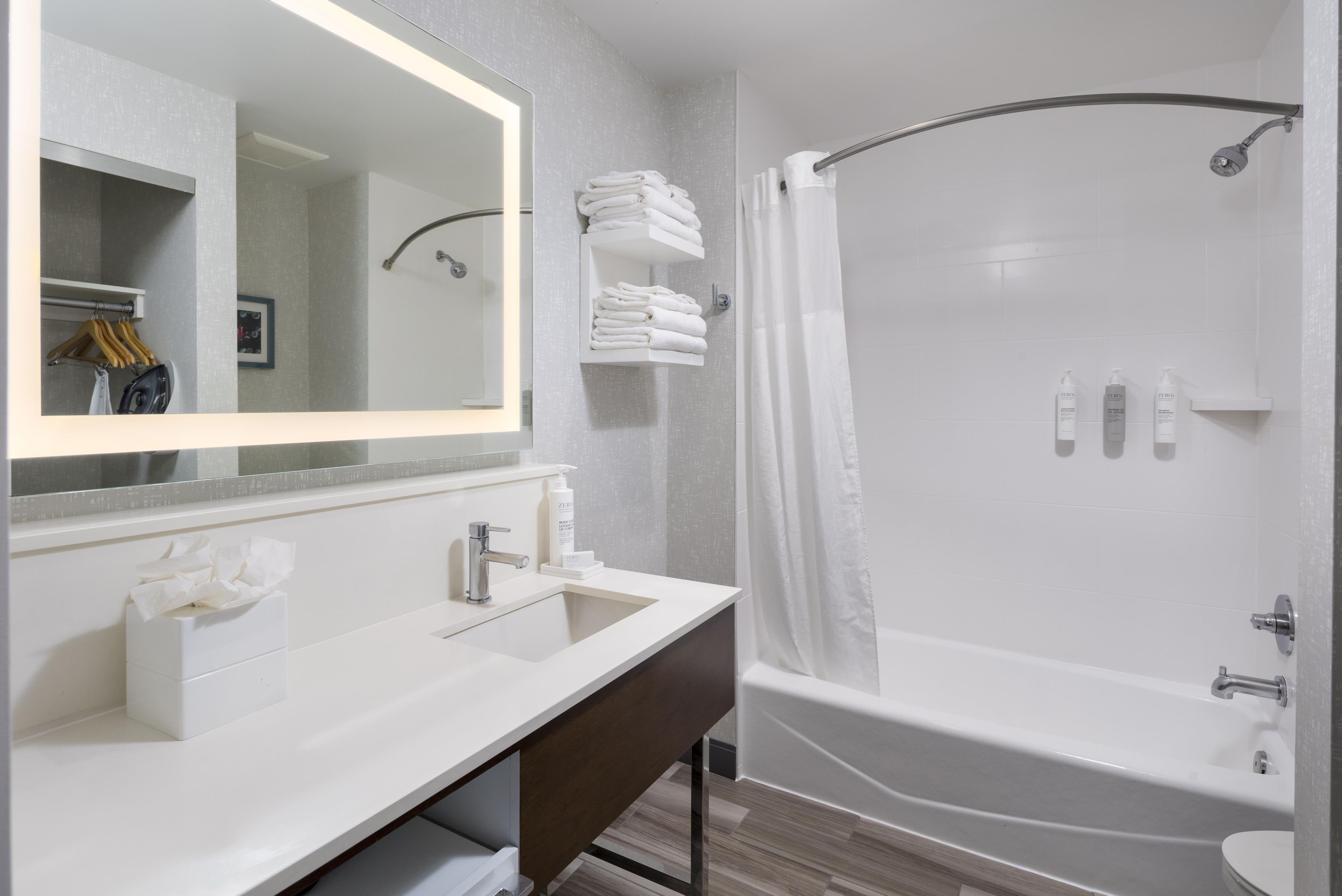 A modern bathroom with a large mirror, a sink, a vanity, a shower with a curved tub, and neatly folded towels on a shelf.