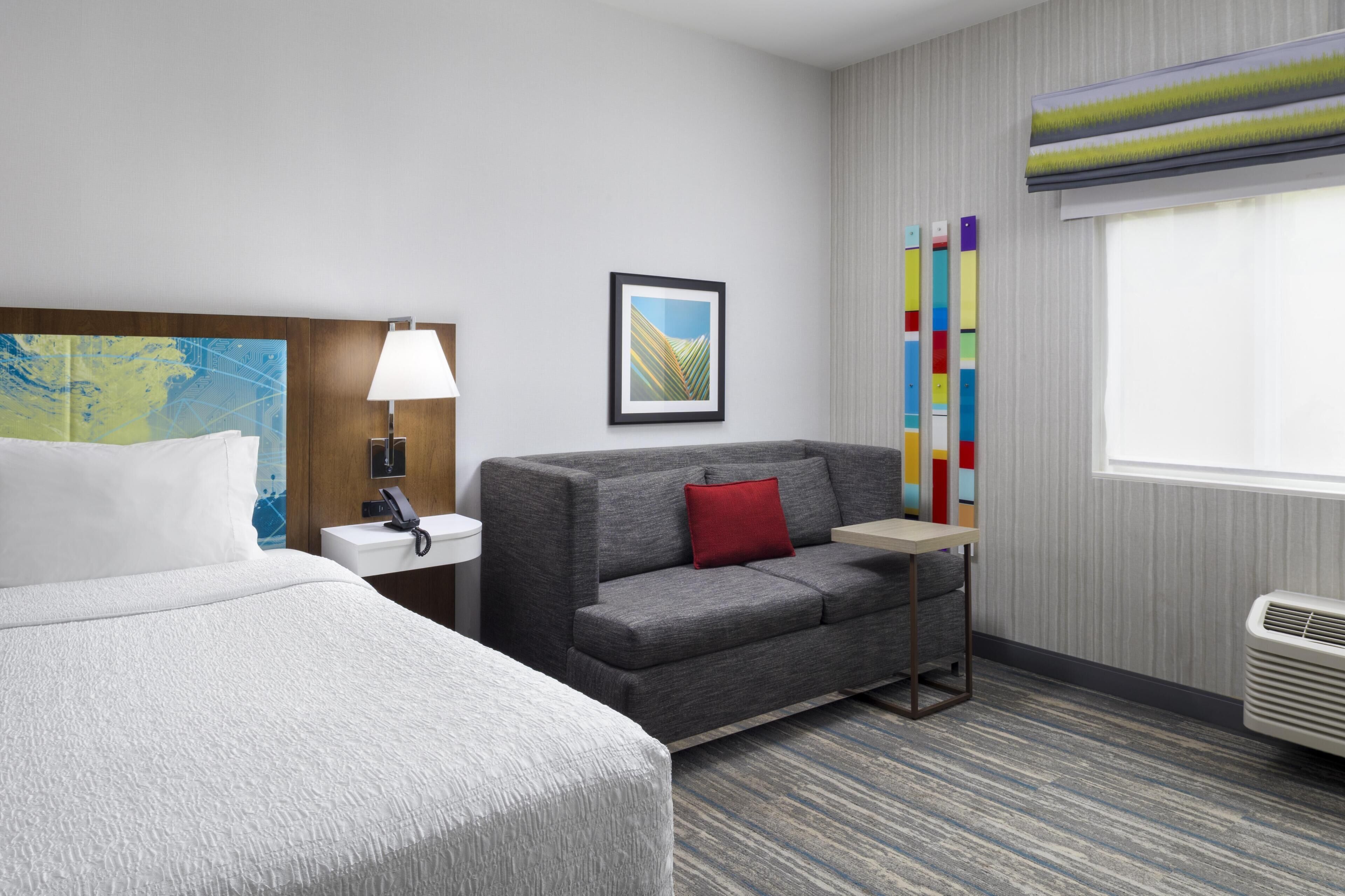Habitación de hotel con cama, sofá, mesita de noche, lámpara, cuadro enmarco, ventana con persianas y radiador.
