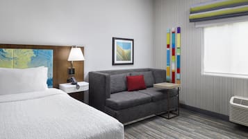 Habitación de hotel con cama, sofá, mesita de noche, lámpara, cuadro enmarco, ventana con persianas y radiador.