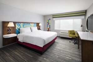 Una habitación de hotel con cama, mesitas de noche, un escritorio con silla, televisión y un ventanal grande.