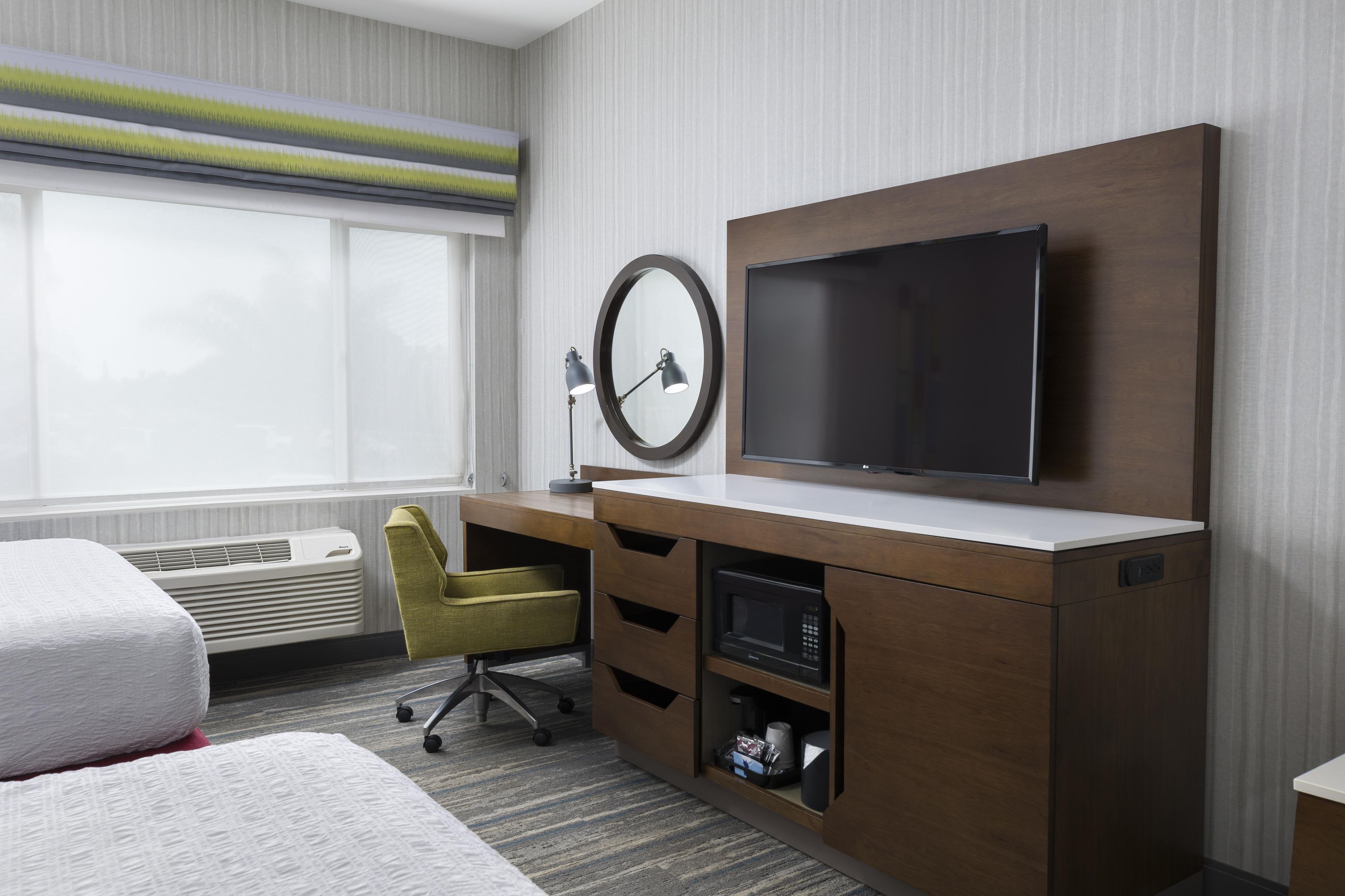Ein Hotelzimmer mit einem Bett, einem Schreibtisch mit Flachbildfernseher, einem Sessel, einer Schreibtischlampe und einem Fenster mit Jalousien.