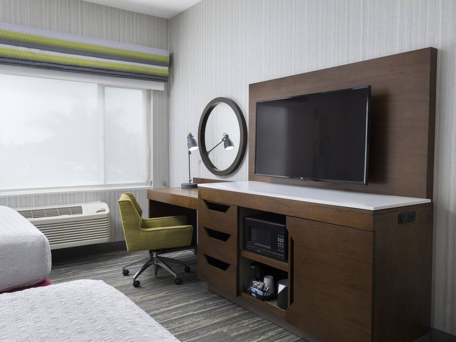 Ein Hotelzimmer mit einem Bett, einem Schreibtisch mit Flachbildfernseher, einem Sessel, einer Schreibtischlampe und einem Fenster mit Jalousien.