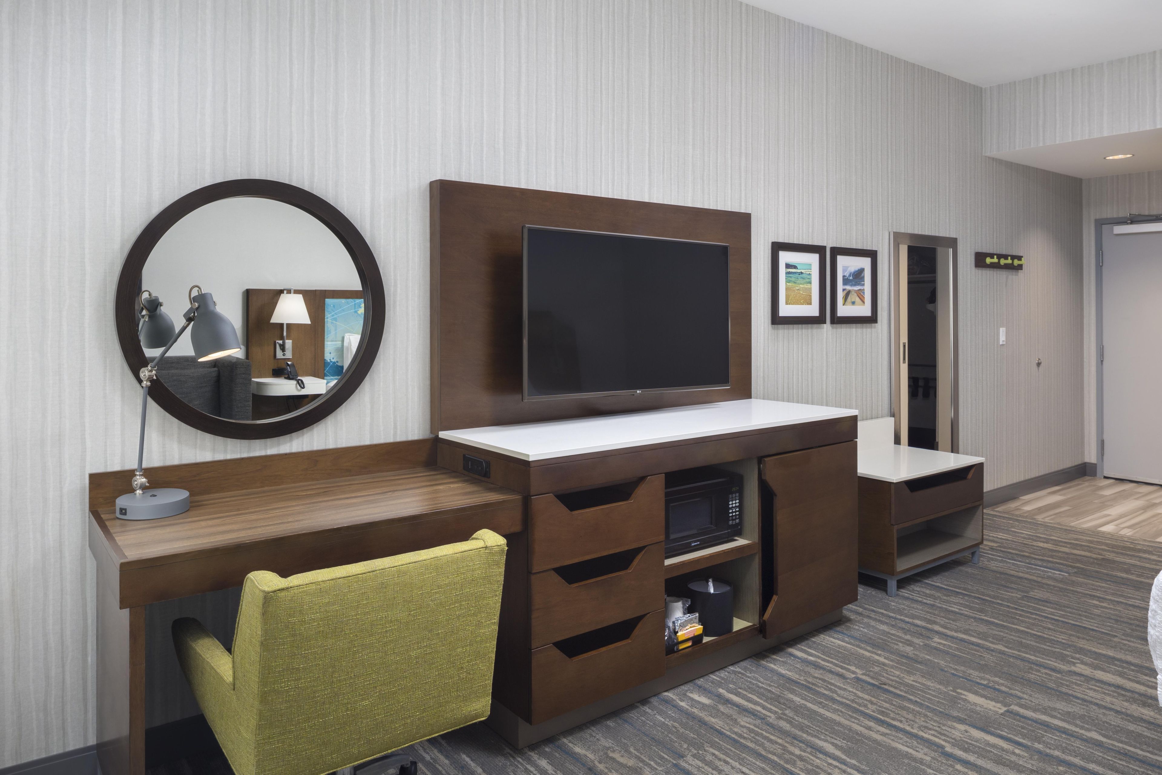 A hotel room with a large flat-screen TV, a round mirror, a desk with a lamp, a chair, and a wardrobe with a microwave.
