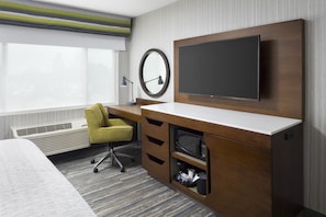 Habitación de hotel con cama, escritorio con televisor de pantalla plana, silla, espejo y ventana con persianas.