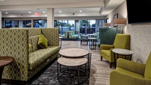 A modern lounge area with patterned sofas, round tables, and a large window offering a view of the outdoors. - Hampton Inn Cardiff Beach Encinitas, CA Near Del Mar (Cardiff-by-the-Sea)