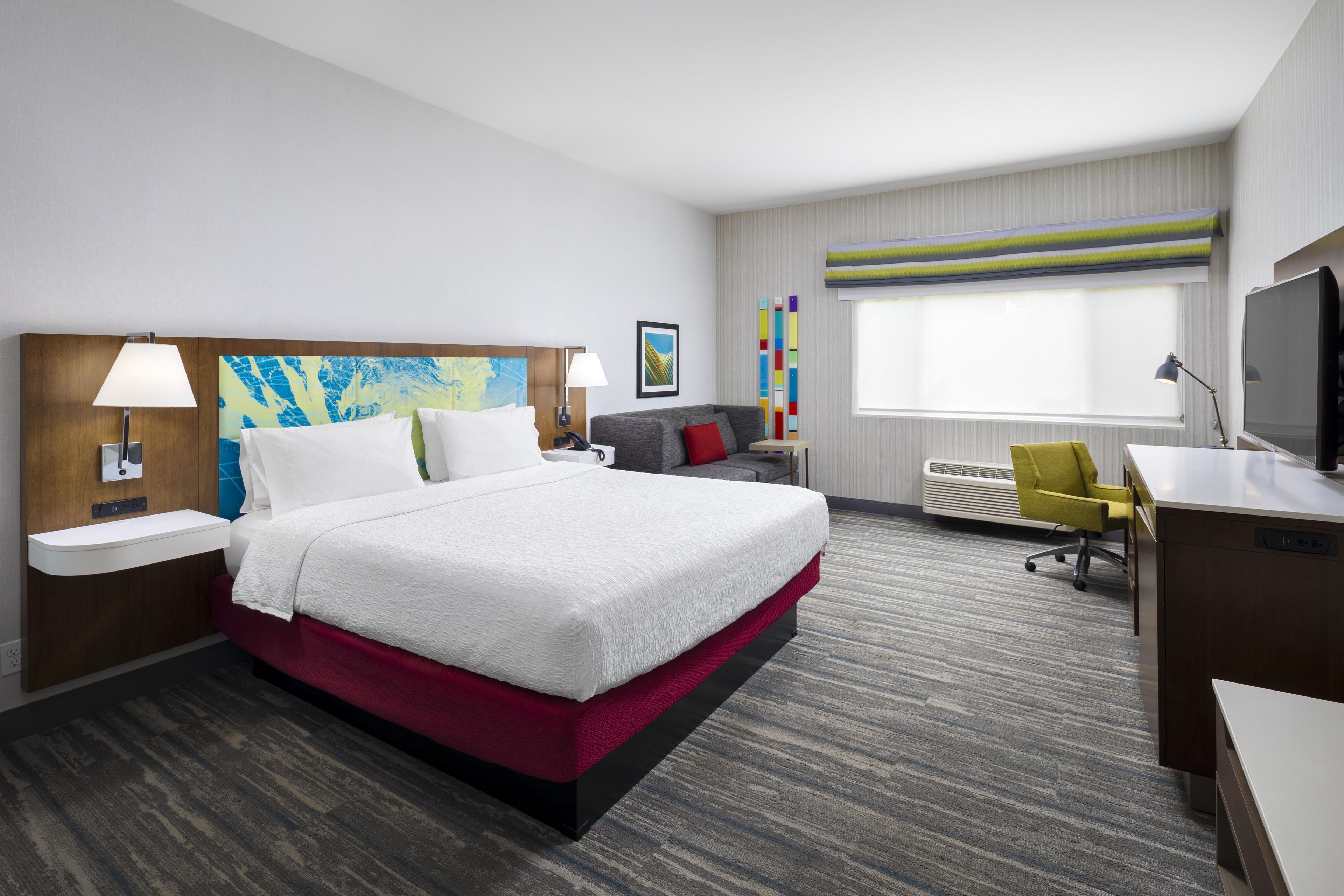 Une chambre d’hôtel comprenant un lit, une table de chevet, une lampe, un tableau, un bureau avec une chaise, un téléviseur et une fenêtre avec des stores.