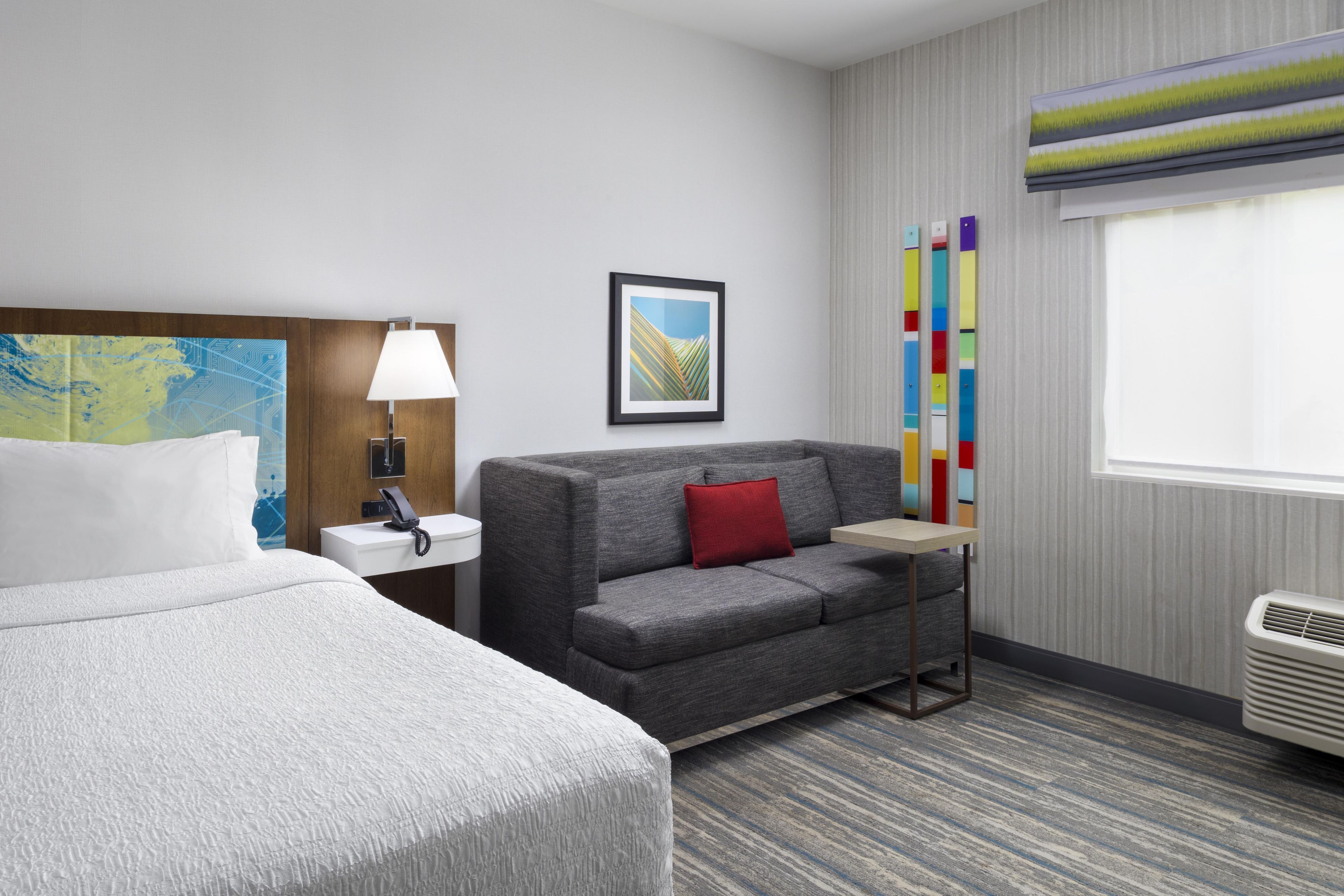 A hotel room with a bed, a sofa, a nightstand, a lamp, a framed picture, a window with blinds, and a radiator.
