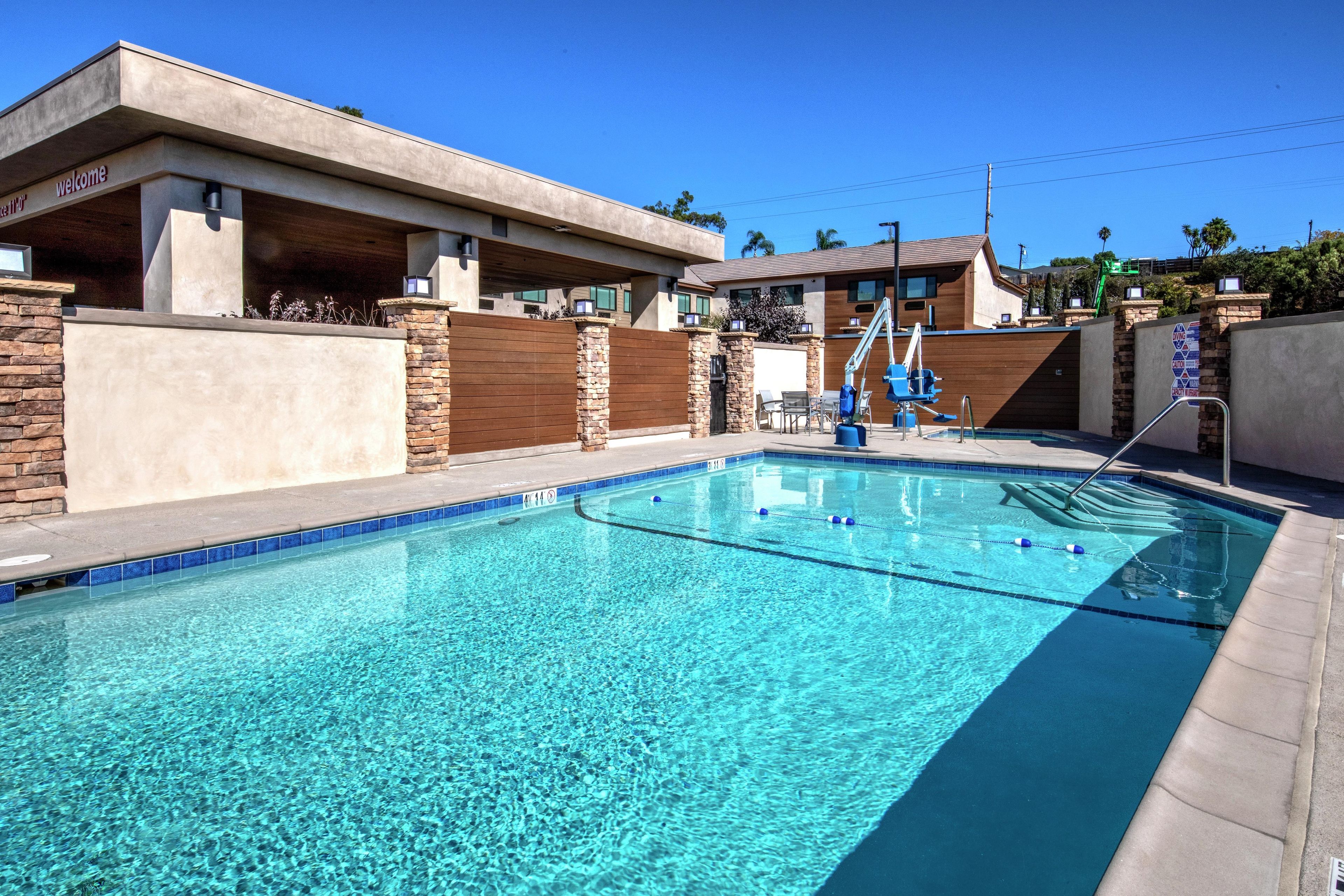 A clear swimming pool with a diving board and lounge chairs, surrounded by a stone wall and modern buildings.
