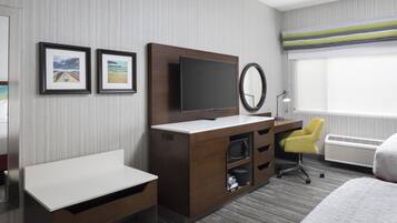 A hotel room with a bed, a desk with a flat-screen TV, a chair, a small table, and a window with a view.