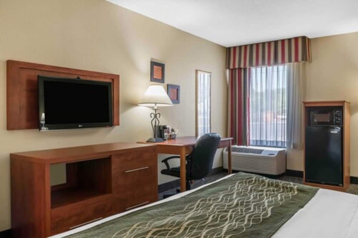 A hotel room with a bed, a desk with a computer, a television, a lamp, a refrigerator, and an air conditioning unit.