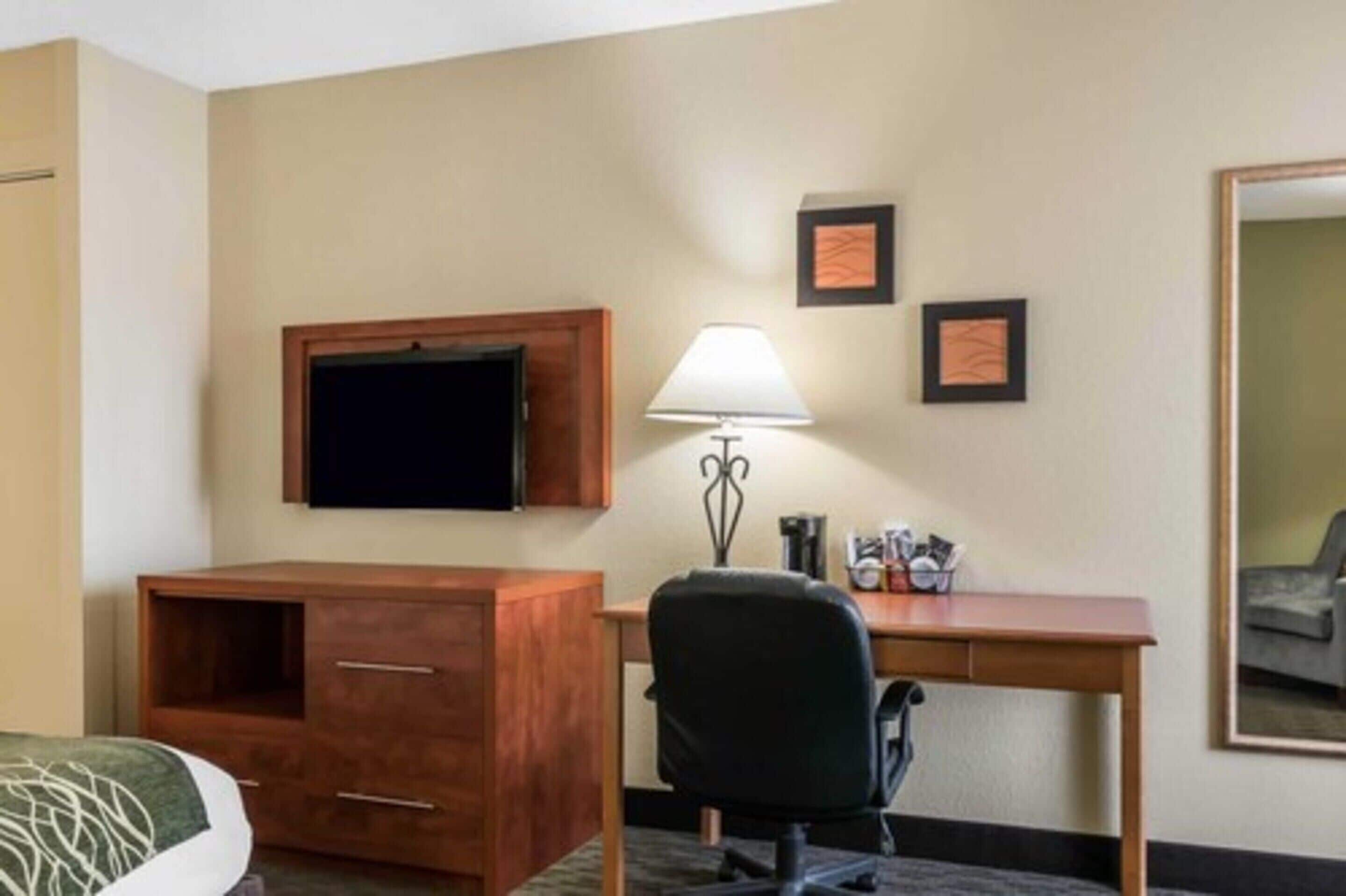 A hotel room with a bed, a desk, a chair, a television, a lamp, and framed certificates on the wall.