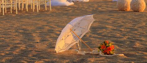 En bröllopslokal på stranden med vita stolar och ett baldakin, ett bord dekorerat med blommor och ett dekorativt parasoll.