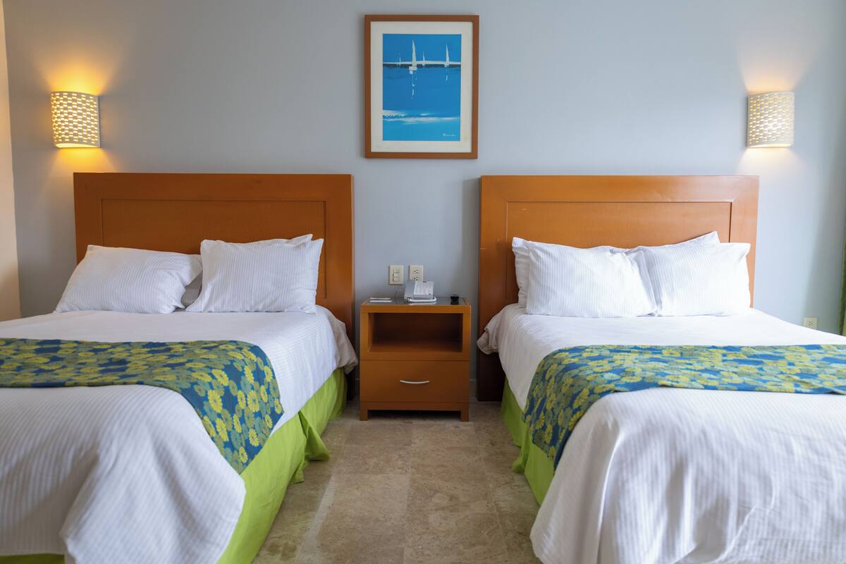 Habitación de hotel con dos camas, cada una con cabecero de madera, ropa blanca y cubrecolchones verdes. Hay una mesita de noche entre las camas, con una lámpara y una foto enmarcada en la pared de arriba.