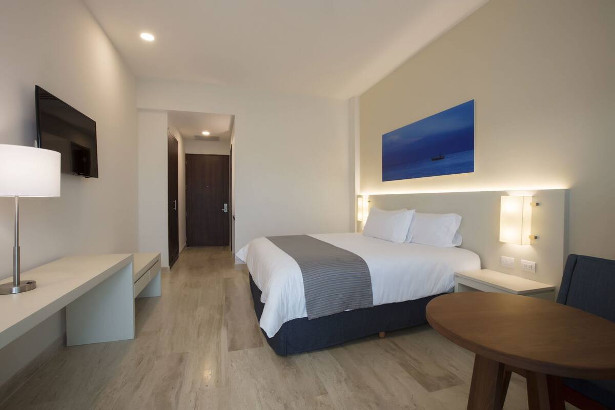 Ein Hotelzimmer mit Bett, Nachttischen, Schreibtisch, Stuhl, einem Gemälde und einer Tür.