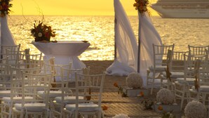 Decoração de casamento com toldo branco, cadeiras e mesa florida, com vista para o mar e um cruzeiro ao fundo.