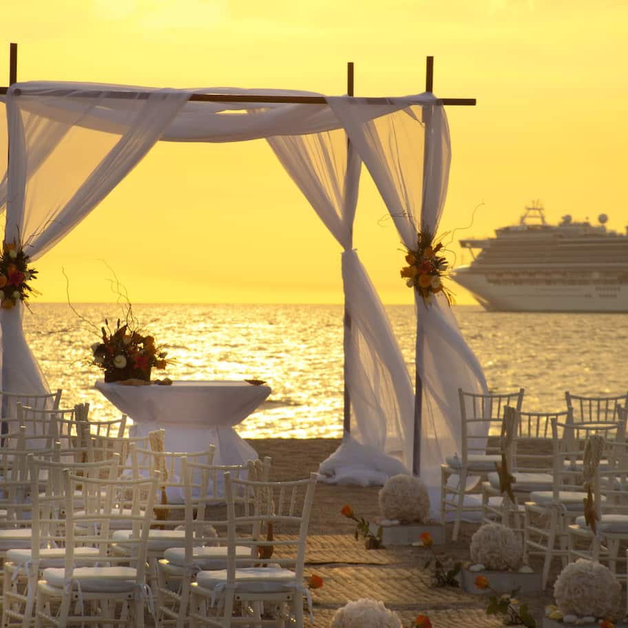 A wedding setup with a white canopy, chairs, and a table with flowers, overlooking the sea with a cruise ship in the background.