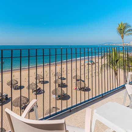 A balcony with a view of a beach, a palm tree, and a fence.